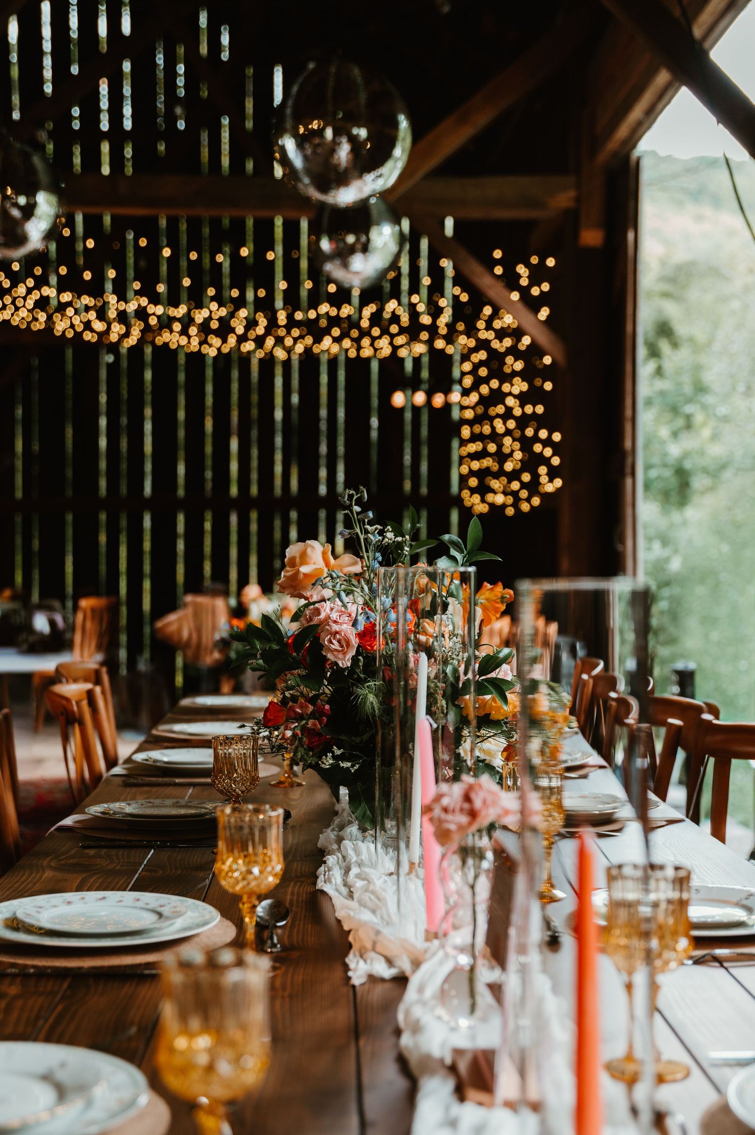 Rustic barn wedding reception table set with string lights, amber glassware and wildflower centerpieces.