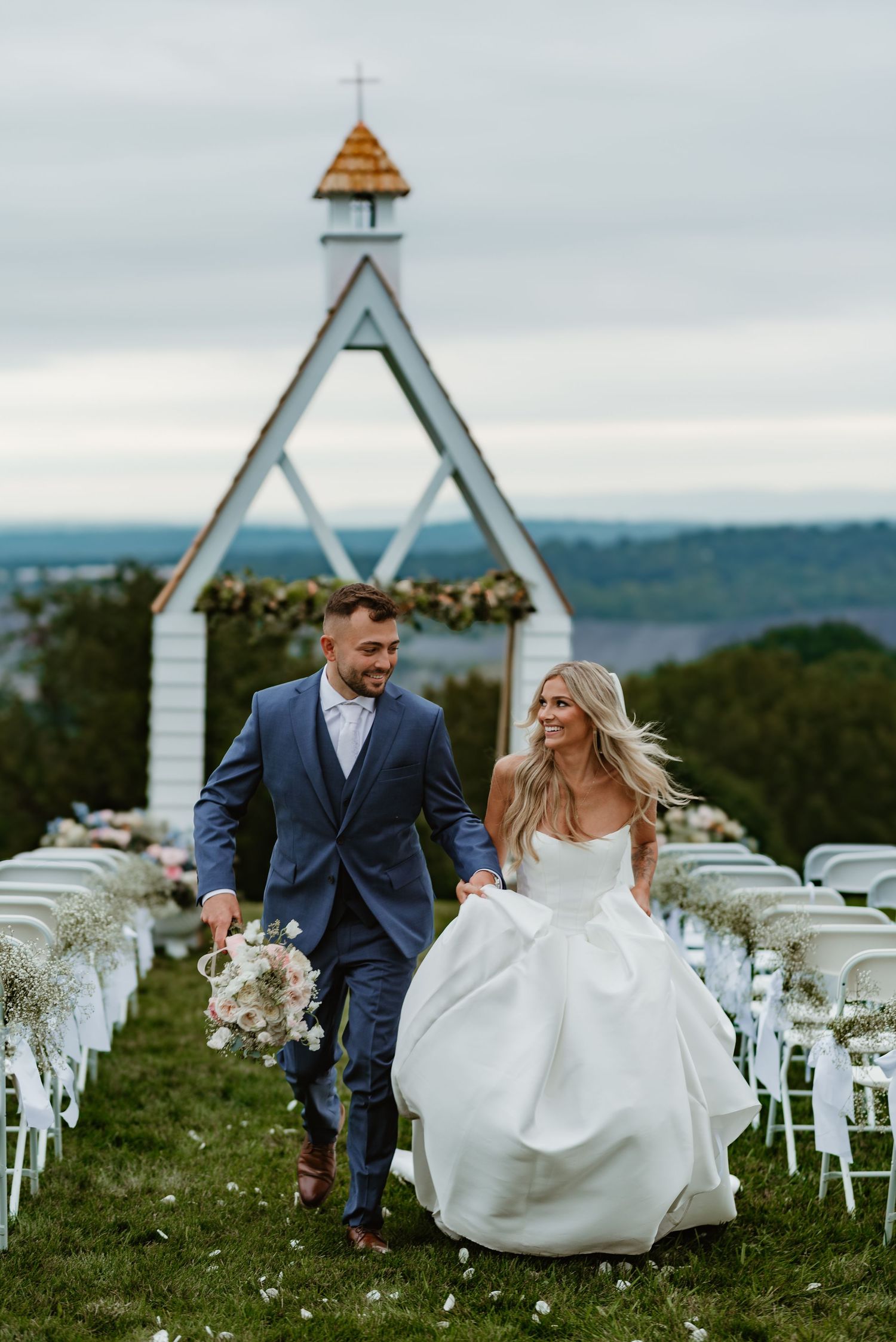 Newlyweds walk through a scenic outdoor wedding venue with white chairs and wildflowers on a cloudy day.