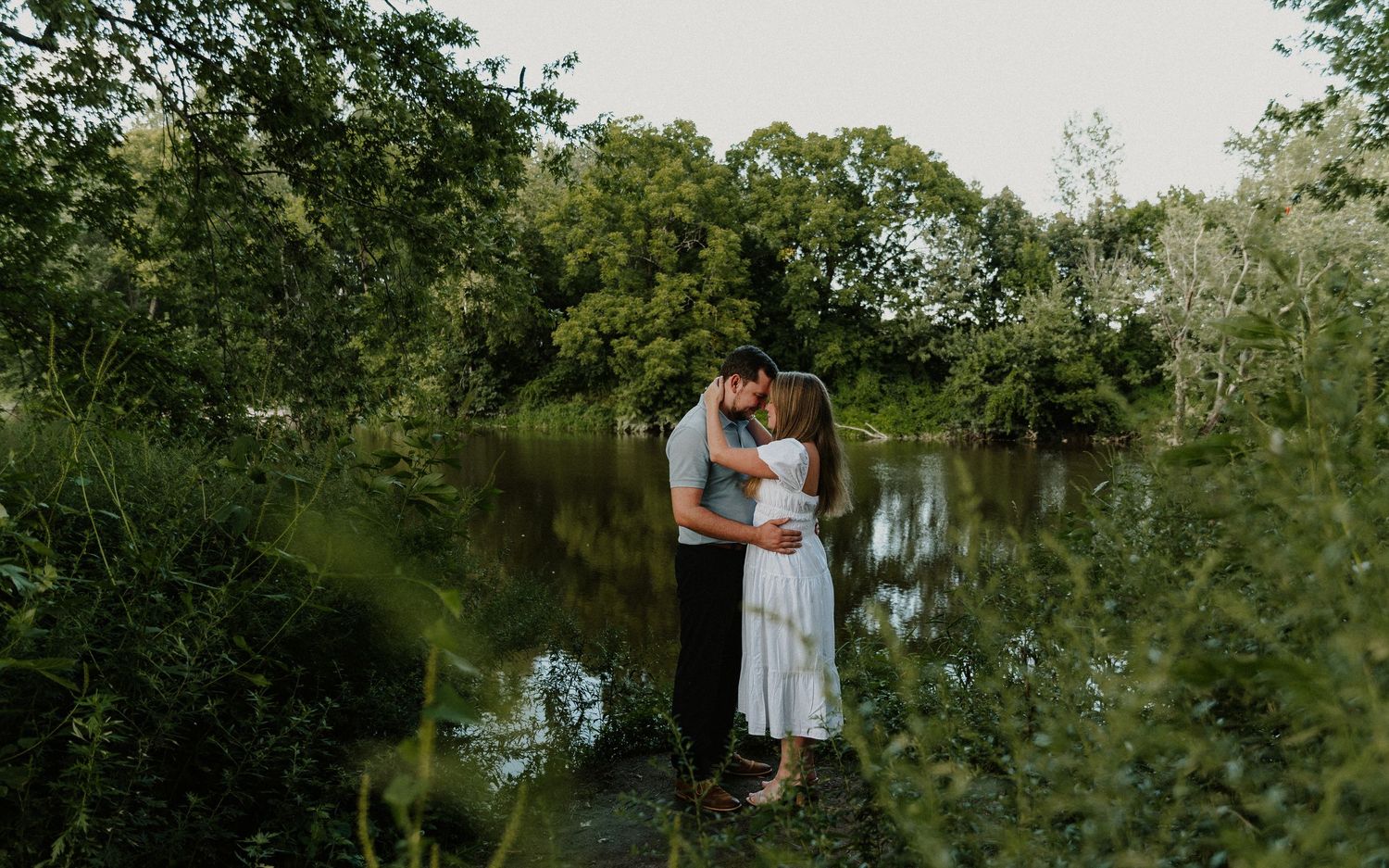 Couple sharing a romantic kiss near a serene pond surrounded by lush green trees and foliage.