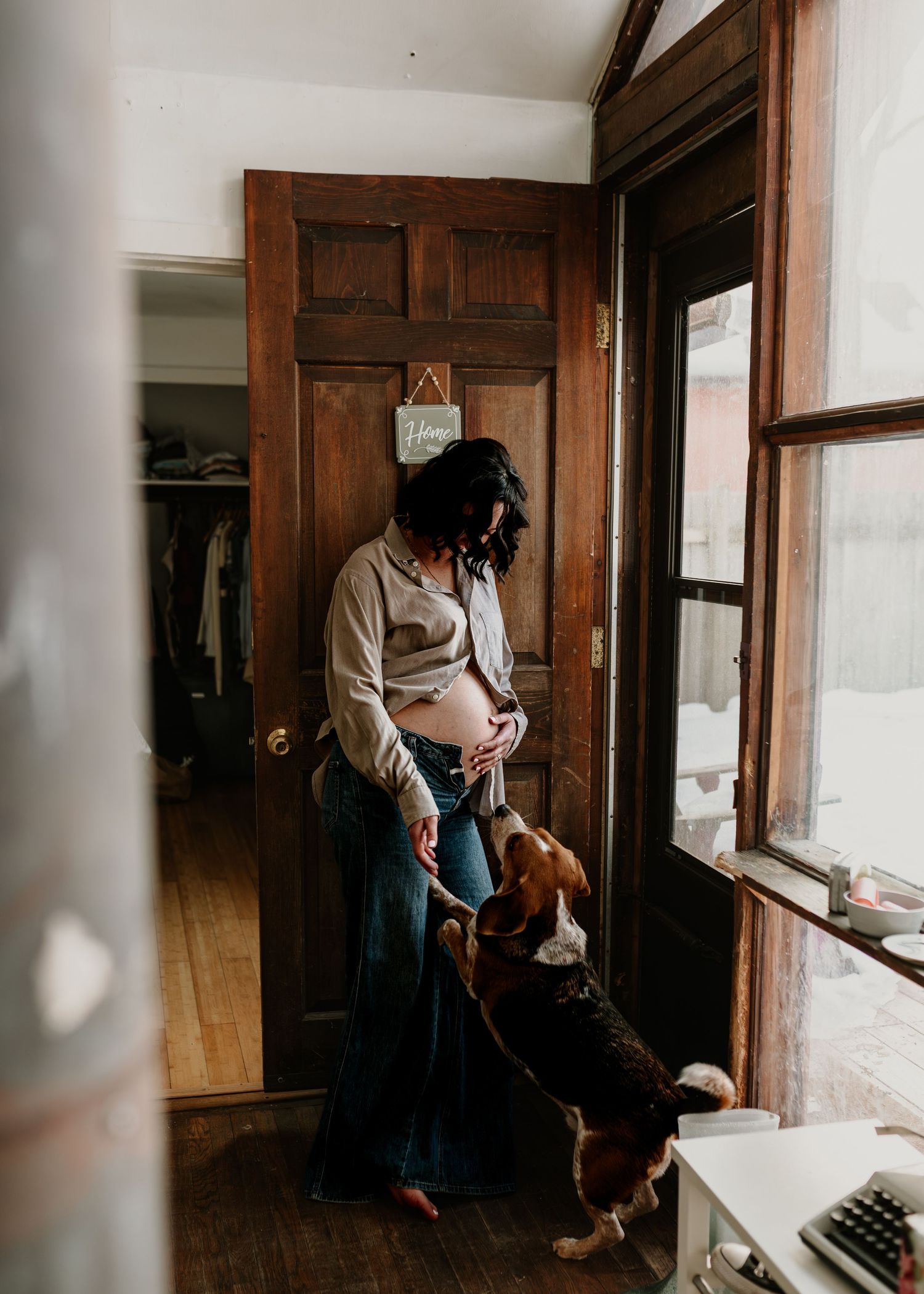 Pregnant woman in jeans cradling baby bump while a beagle dog nuzzles her near a wooden door indoors.