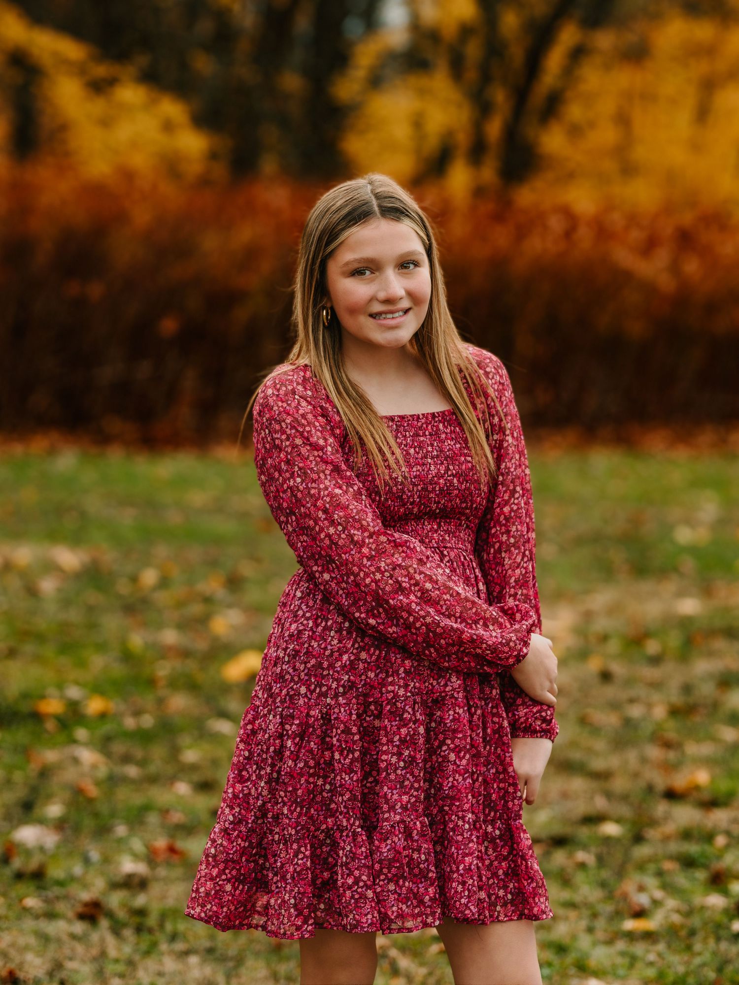 A person wearing a burgundy floral dress poses in a fall setting with autumn leaves on the ground.