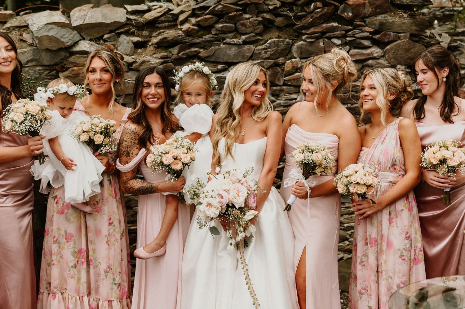 Wedding party in pink and mauve dresses poses together holding white and pink floral bouquets against stone wall backdrop.