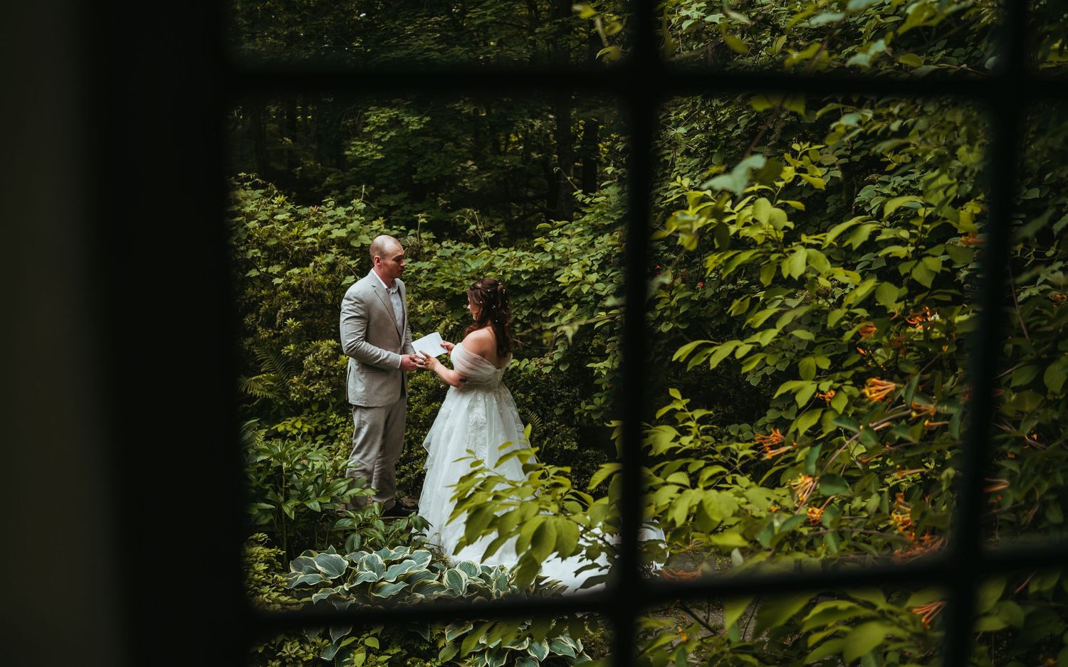 A couple in wedding attire walks hand in hand through a lush green forest path.