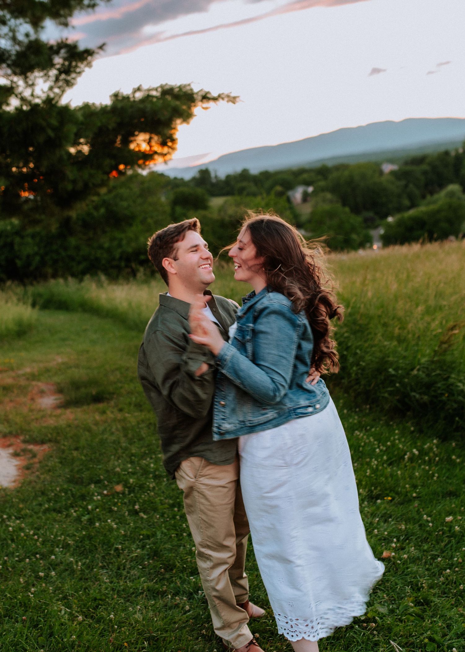 Couple embraces and laughs together in a green meadow at sunset with mountain views in the background.