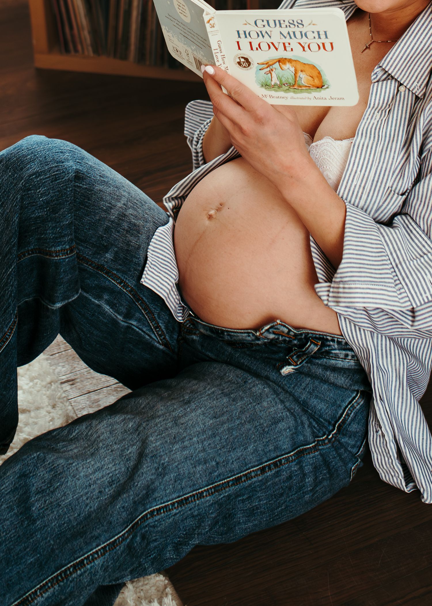 Pregnant woman sitting on floor reading Guess How Much I Love You book with belly exposed wearing jeans.