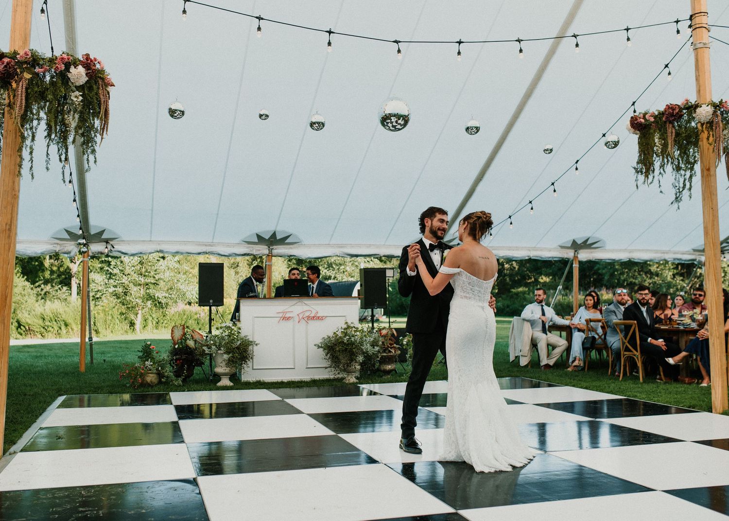 Bride and groom share first dance on black and white checkered floor under elegant outdoor wedding tent.