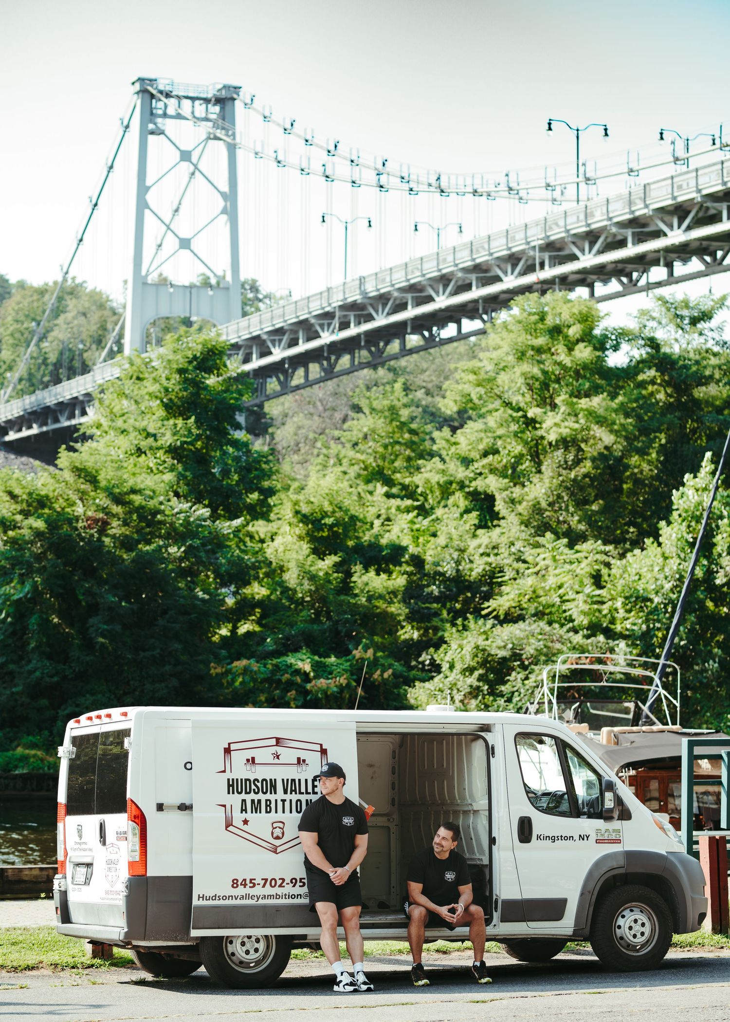 Mobile coffee truck parked under a large suspension bridge with green trees and foliage in the background.