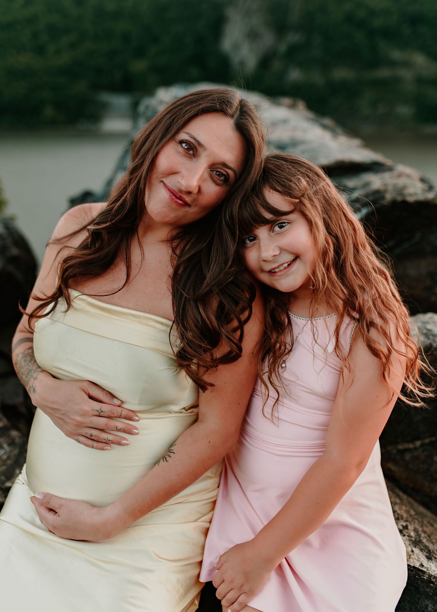 Pregnant woman in yellow dress poses with young girl in pink dress on rocky outdoor setting.