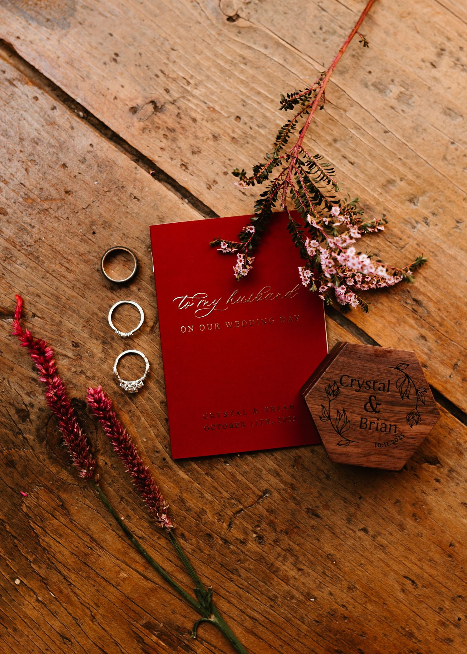 Wedding rings and a red invitation card with dried flowers rest on a rustic wooden surface.