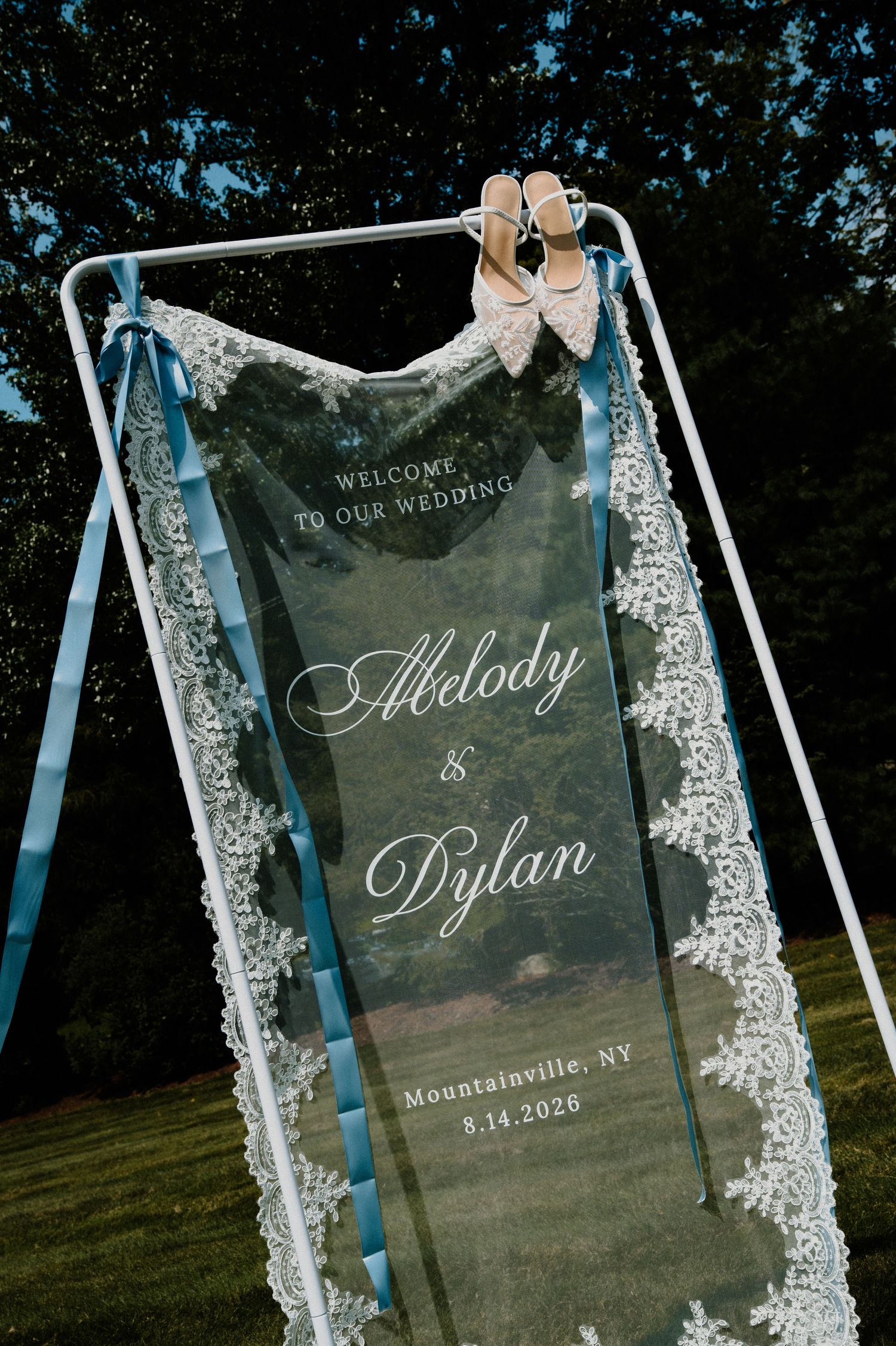 Elegant acrylic wedding welcome sign for Melody and Dylan, Mountainville NY, adorned with lace veil and bridal heels outdoors.