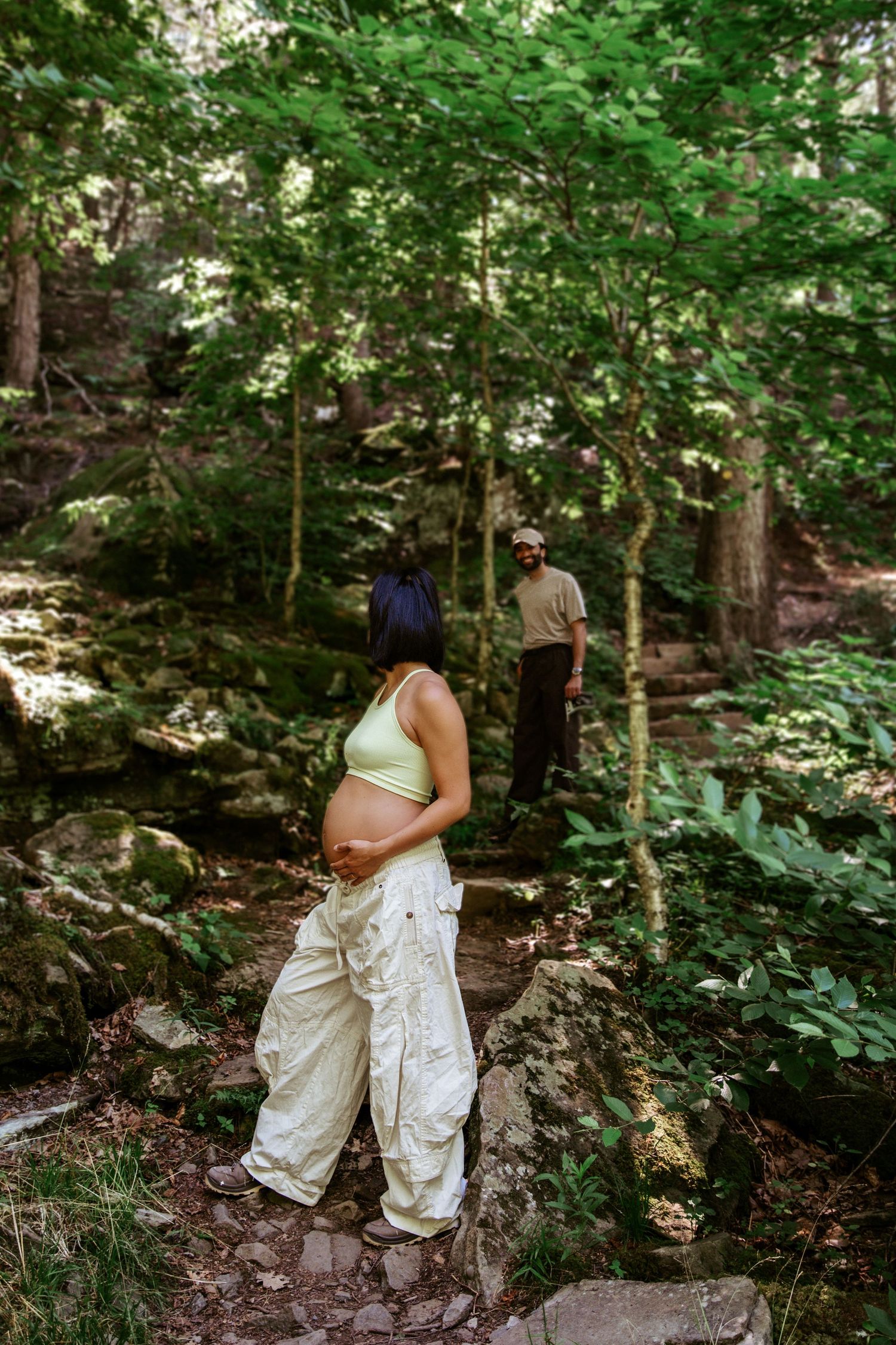 Pregnant woman in green crop top and white cargo pants stands on rocks in lush forest, man visible in background.