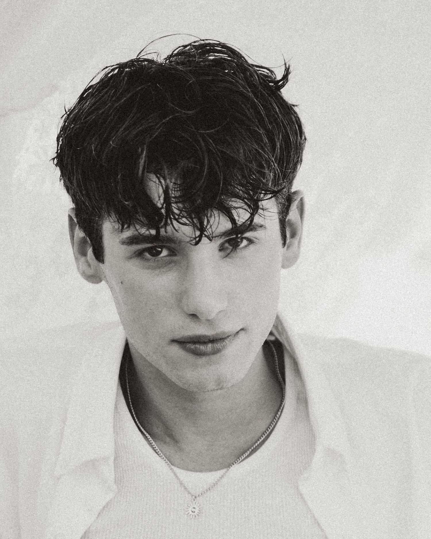 Black and white portrait of a young man with dark wavy hair wearing a white shirt and delicate necklace.