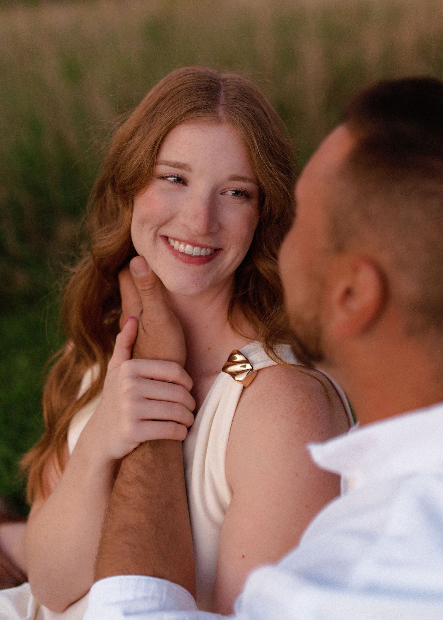Smiling woman with long wavy red hair gazes warmly at a man as he gently holds her hand to her shoulder in a sunlit grassy field.