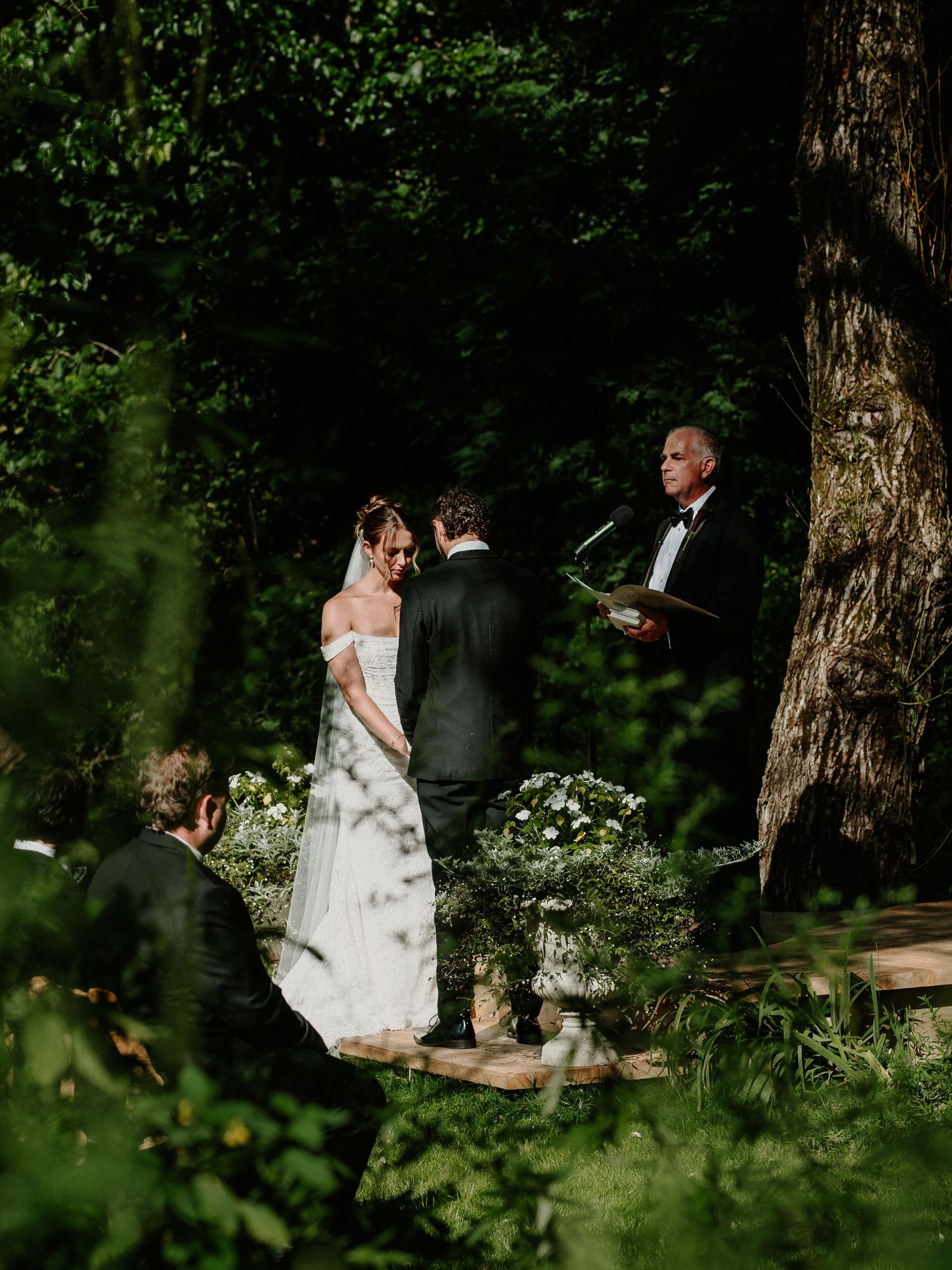 Bride and groom exchange vows in a lush forest ceremony, officiant reading from a book beside a large tree.