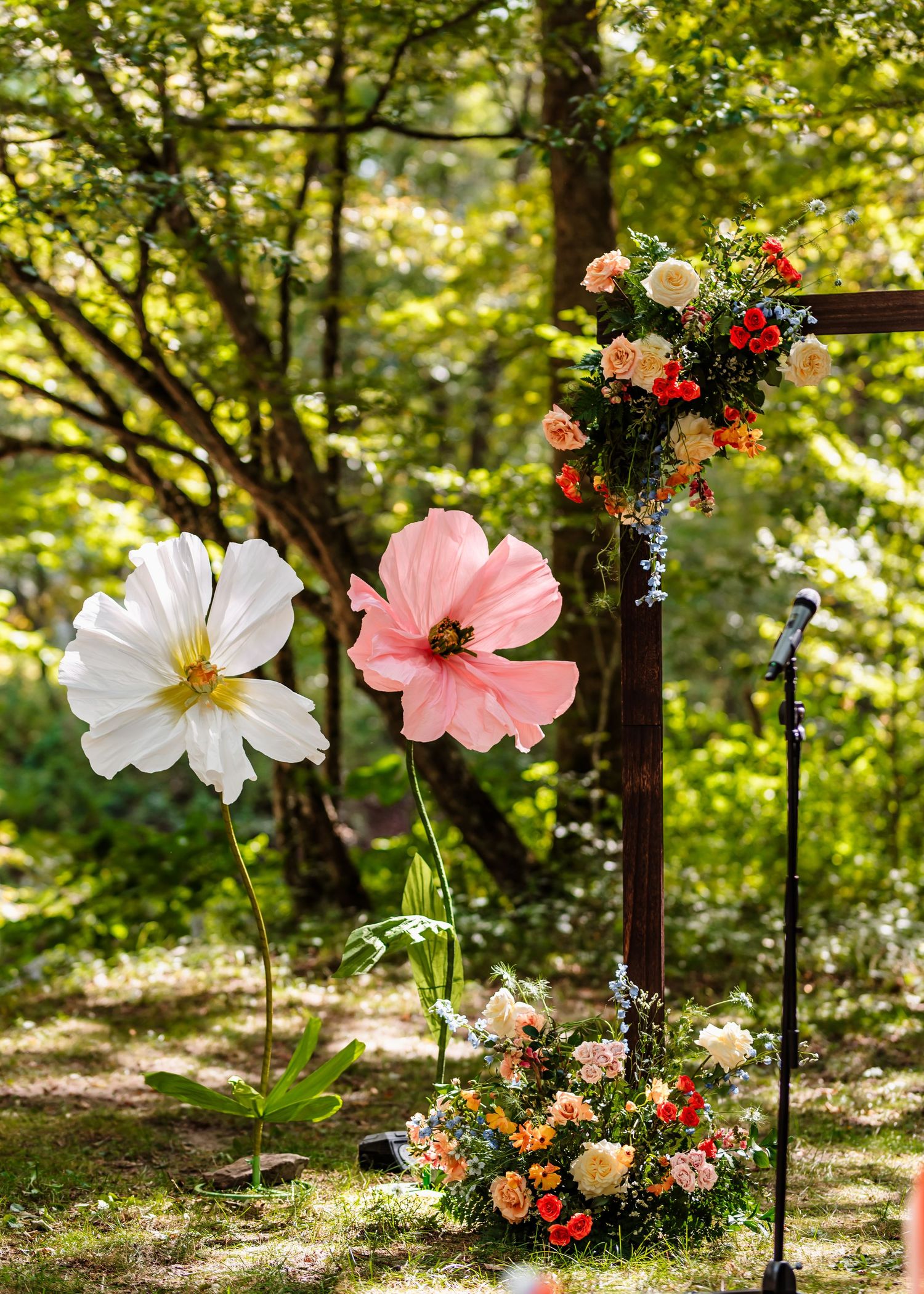 Delicate pink and white cosmos flowers bloom against a sunlit forest backdrop with decorative garden stakes.