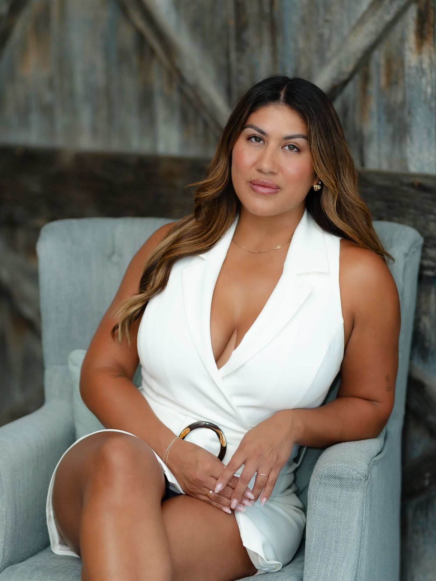 Woman with long highlighted hair in a white sleeveless wrap dress sits in a gray tufted armchair against a rustic weathered wood wall.