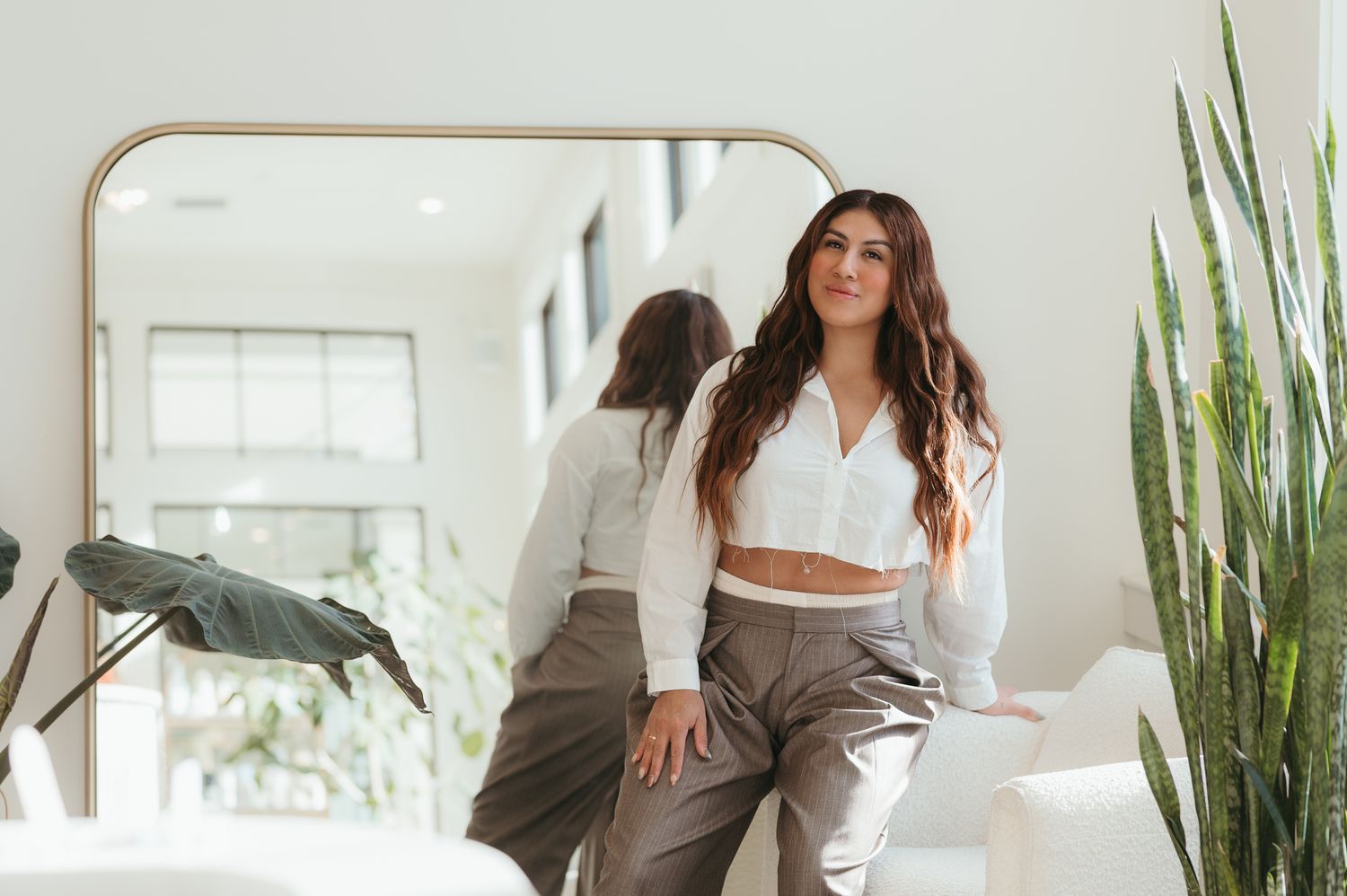 Professional wearing brown blazer stands in modern bright office space with indoor plants and white decor.