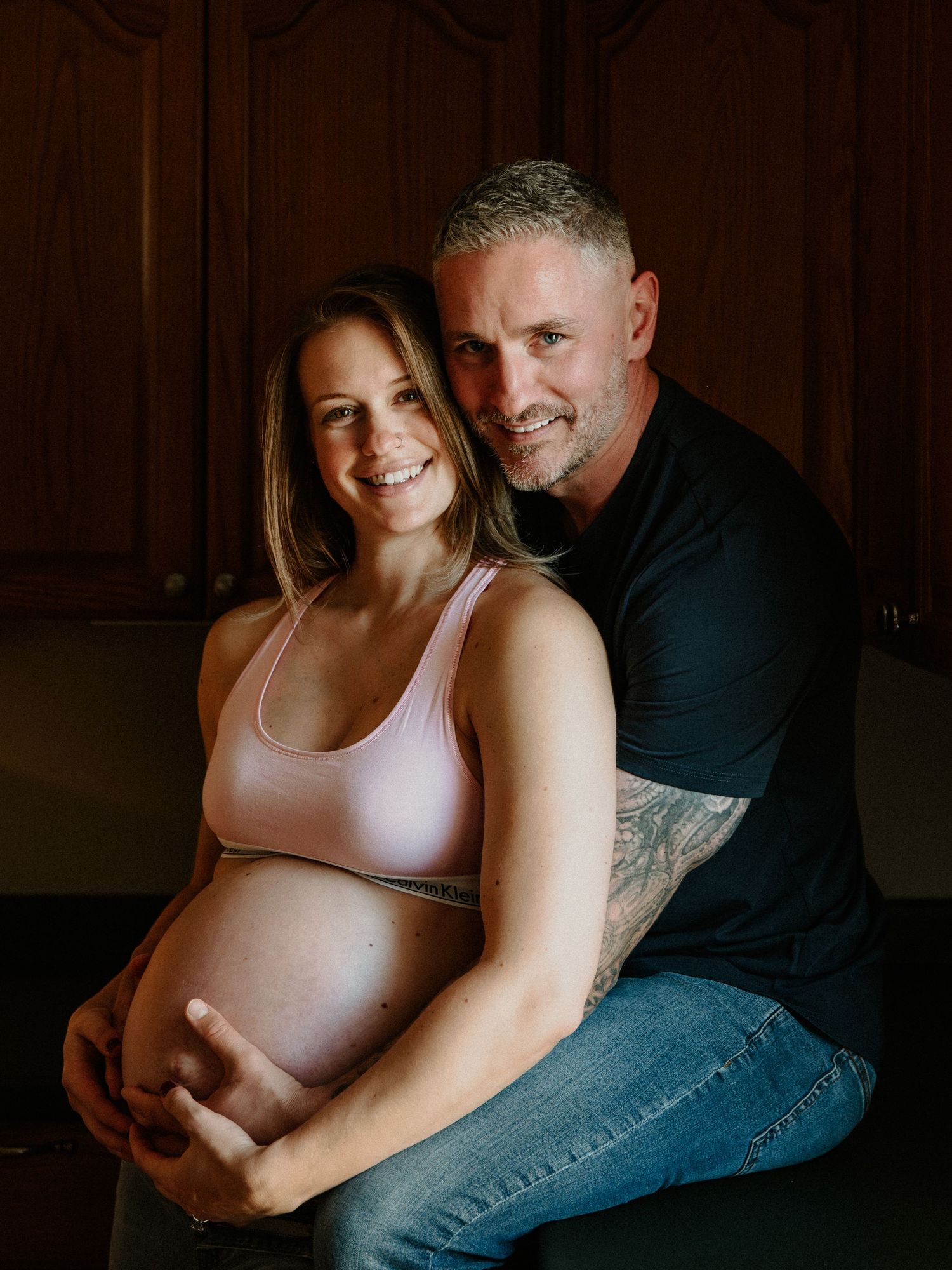Intimate maternity portrait showing a couple embracing in a dimly lit room with wooden paneled walls.