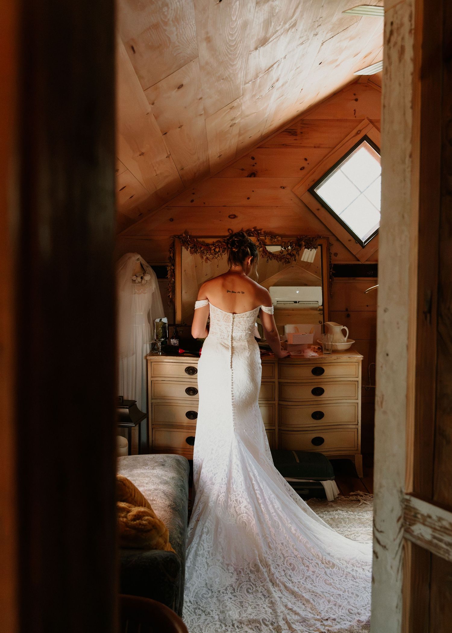 Bride in lace off-shoulder wedding gown with train stands before dresser in rustic wood cabin bridal suite.