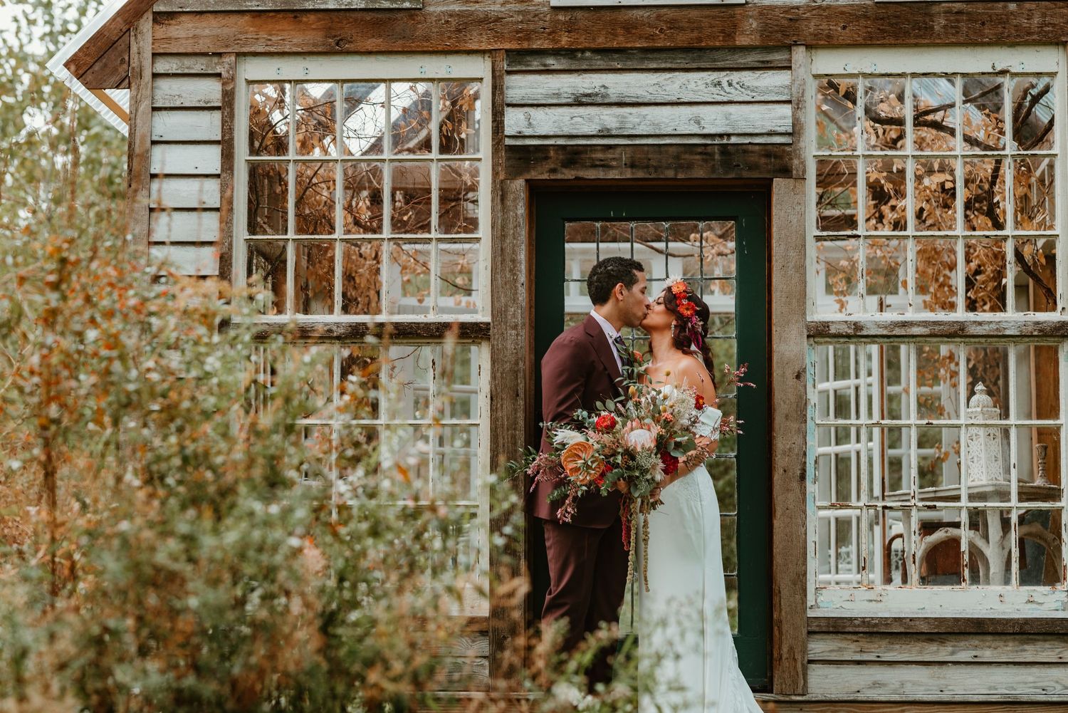 A romantic wedding couple stands in a doorway of a rustic greenhouse surrounded by autumn foliage and vintage windows.