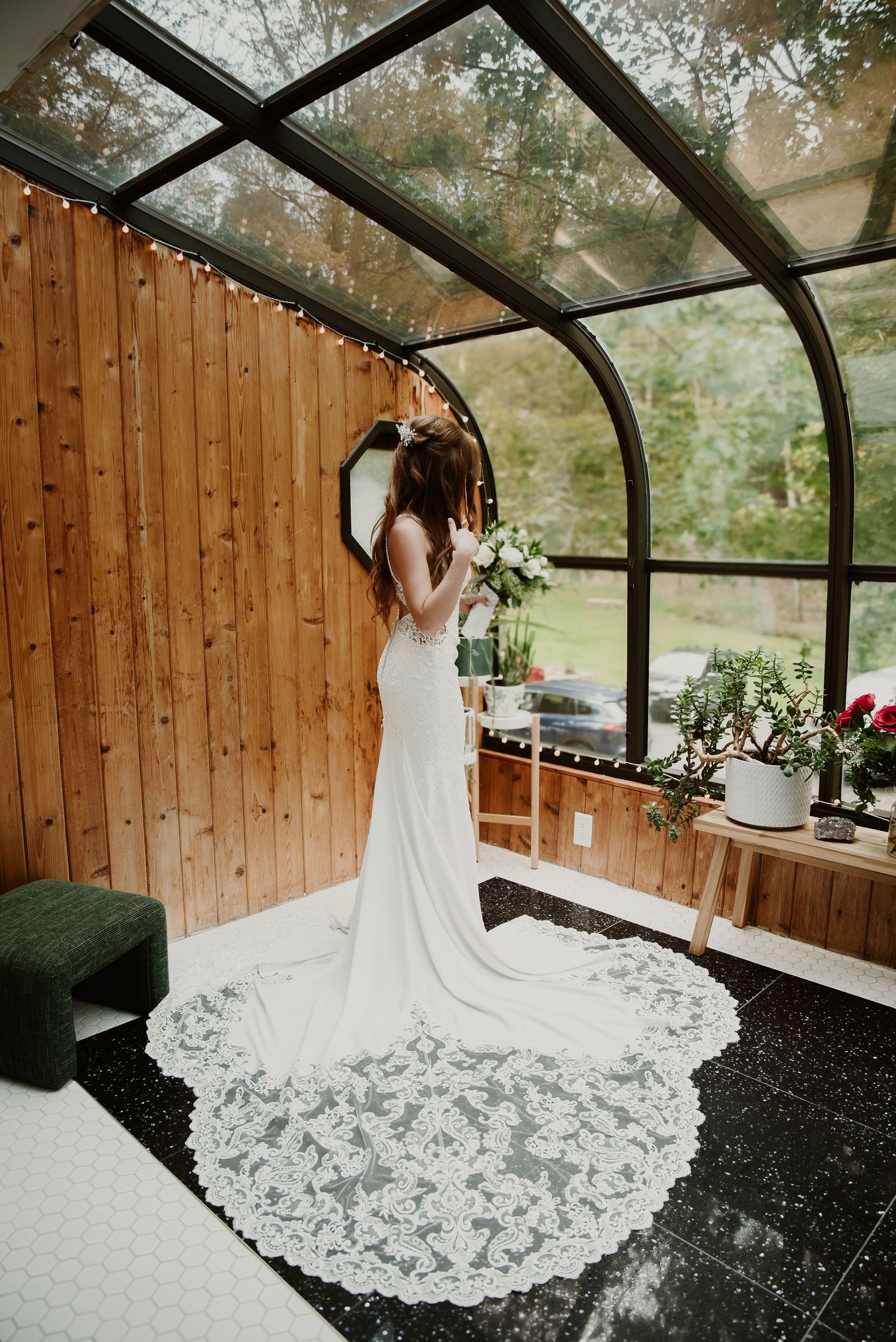 Bride in a flowing lace wedding gown with long train standing in a glass-ceiling greenhouse.