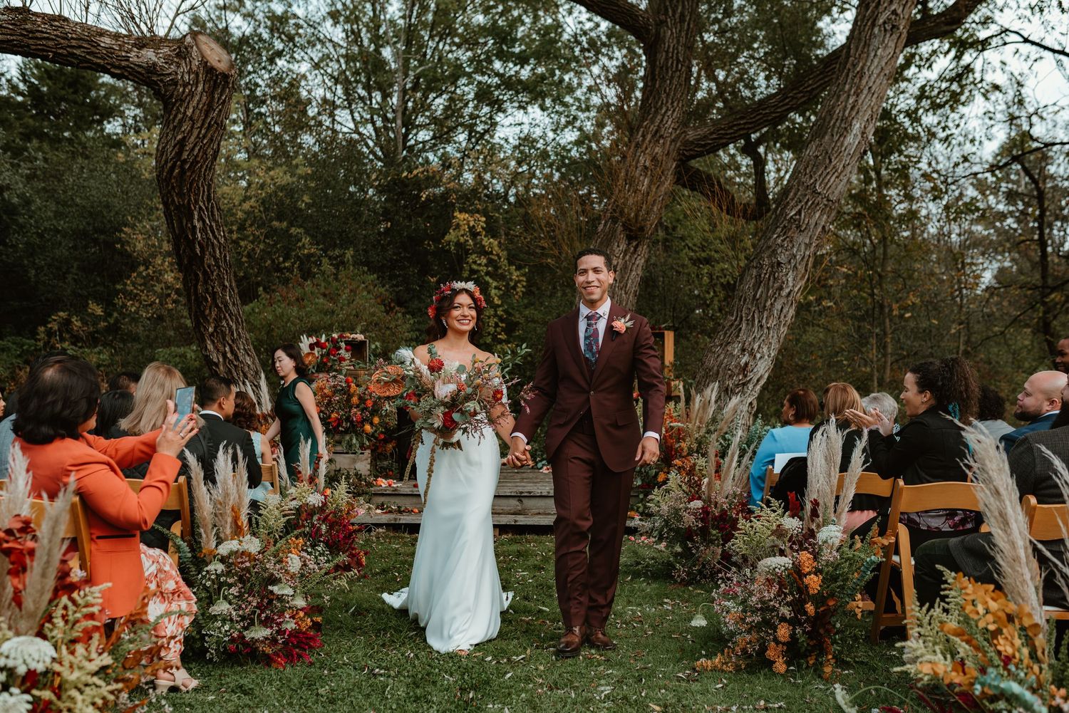 A couple walks down the aisle at an outdoor autumn wedding ceremony with rustic floral arrangements and guests seated.