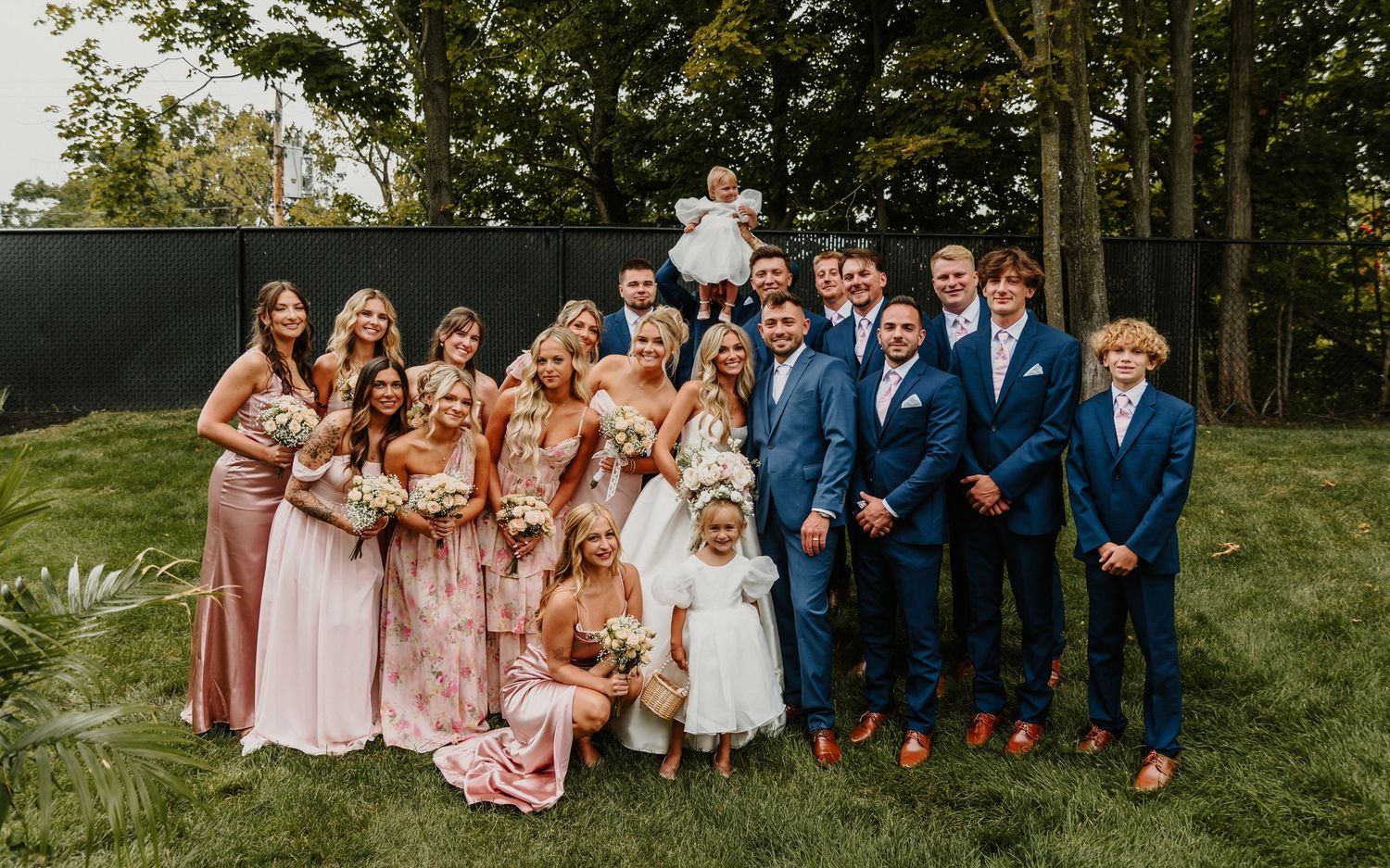 Wedding party poses together outdoors in coordinated pink dresses and navy suits for formal group photos.