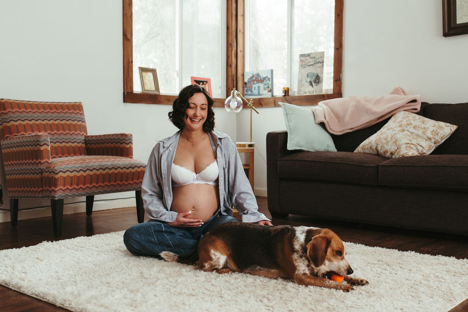Pregnant woman sitting cross-legged on white rug with Cavalier King Charles Spaniel dog in cozy living room.