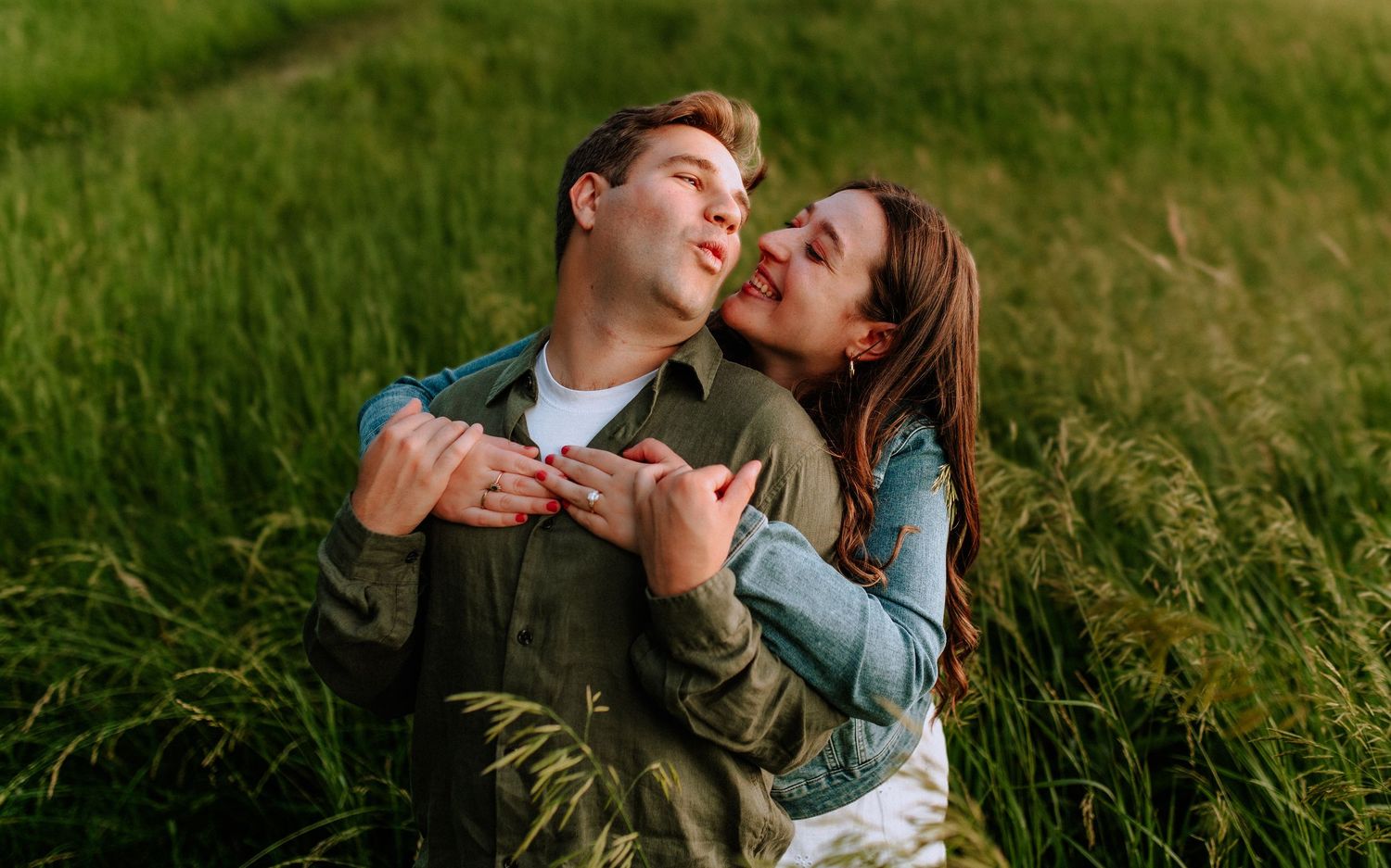 A couple embraces romantically in a lush green field, the woman kissing the man's cheek as he smiles.