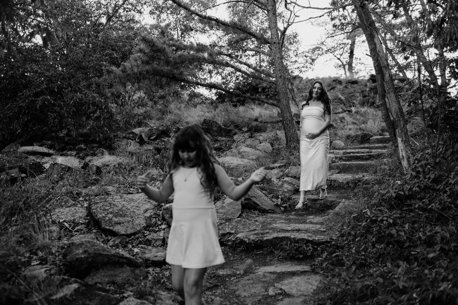 Black and white photo of a young girl in a dress walking down rocky stone steps in a wooded area, with a woman in a long dress behind her.
