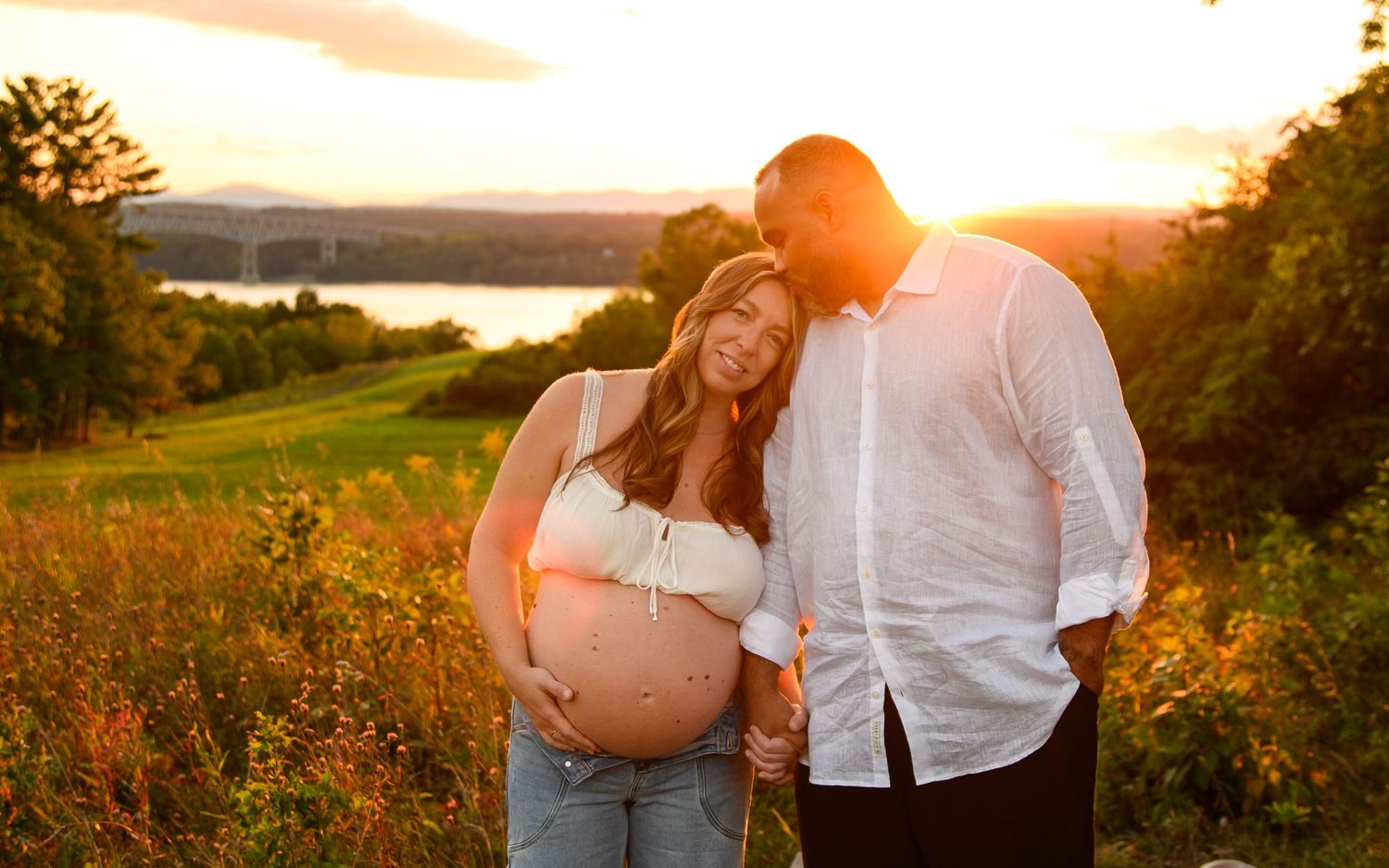 A couple embraces in a golden sunset field with mountains and water in the background.