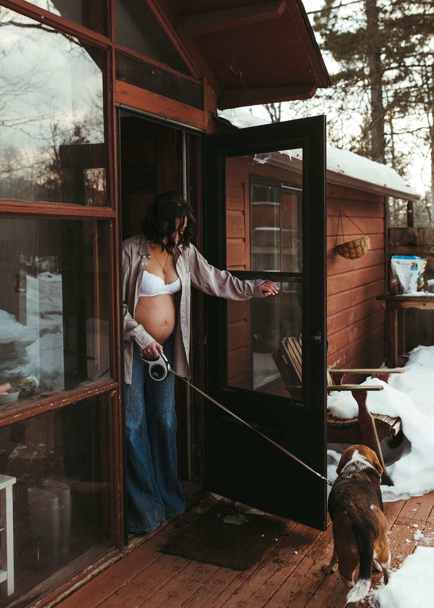 Pregnant woman in white bra and jeans holds dog leash while opening cabin door in snowy winter.