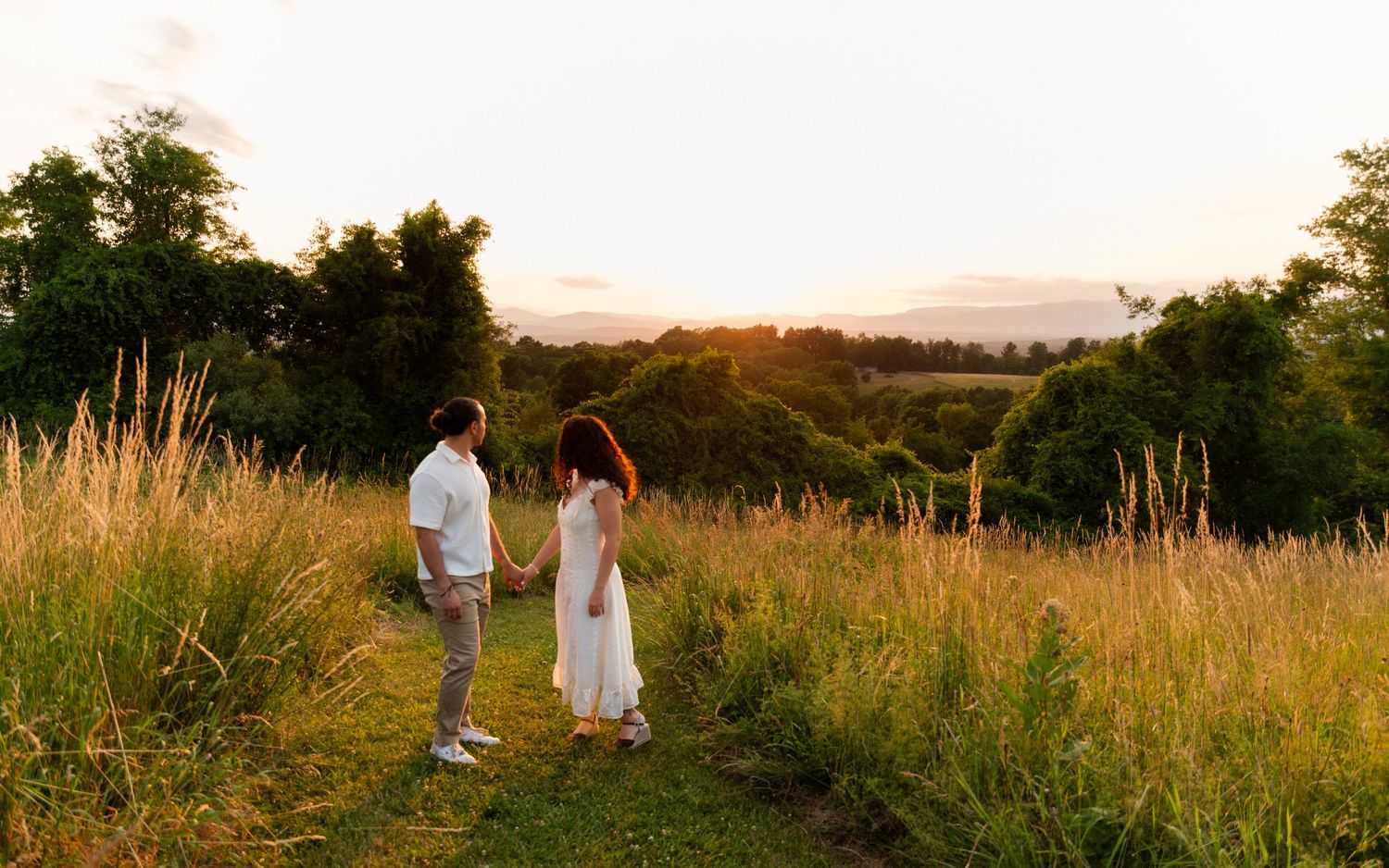Couple holding hands in a golden sunset meadow surrounded by lush green trees and tall grass.