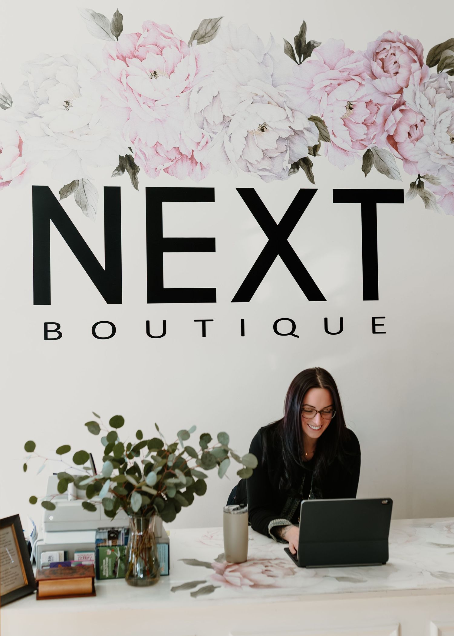 People working at desks in a modern boutique office space with pink floral wall art and potted plants.