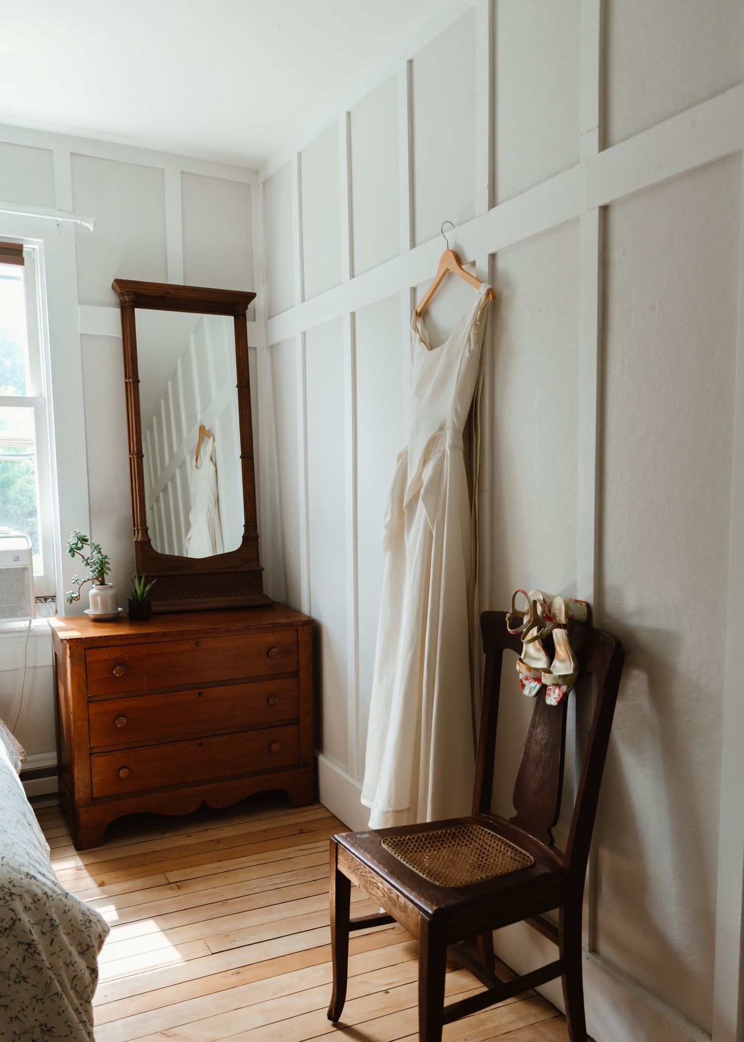 A bright white bedroom with wood floors features vintage dressers, chairs, and white board and batten wall paneling.