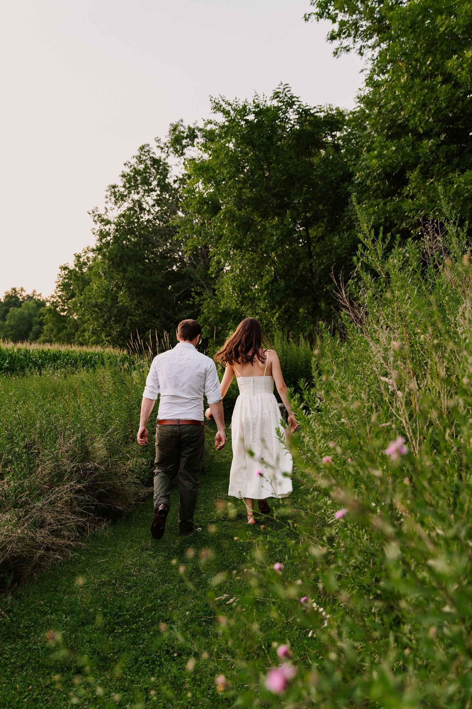Couple walking hand in hand through a lush green field path, woman in white dress, man in white shirt and dark pants.