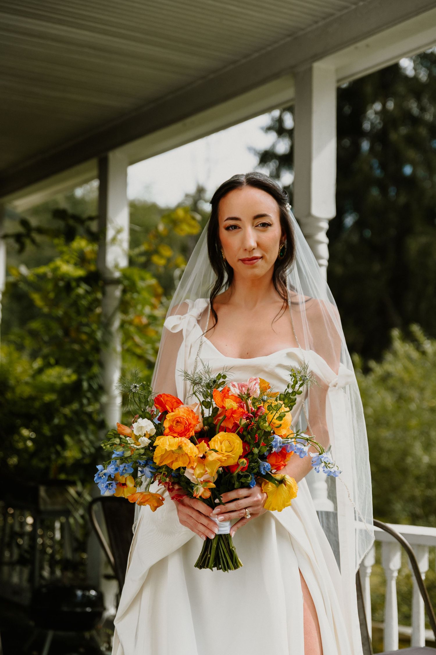 A bride in a white wedding dress stands on a porch holding a vibrant bouquet of orange, yellow and blue flowers.