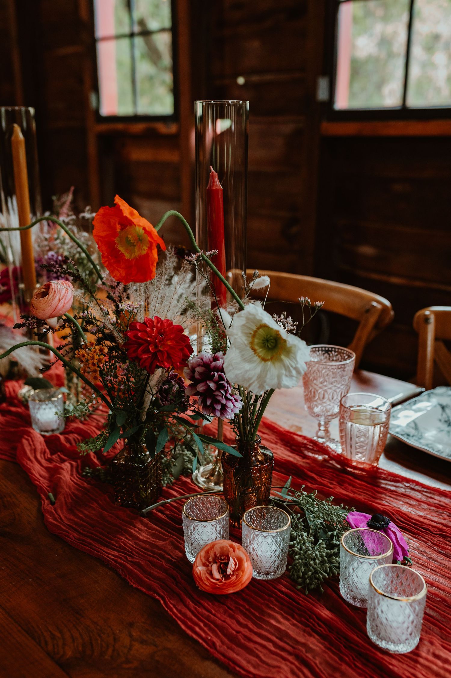 Rustic wedding table decor with colorful wildflowers in vases and glass candle holders on a red tablecloth.