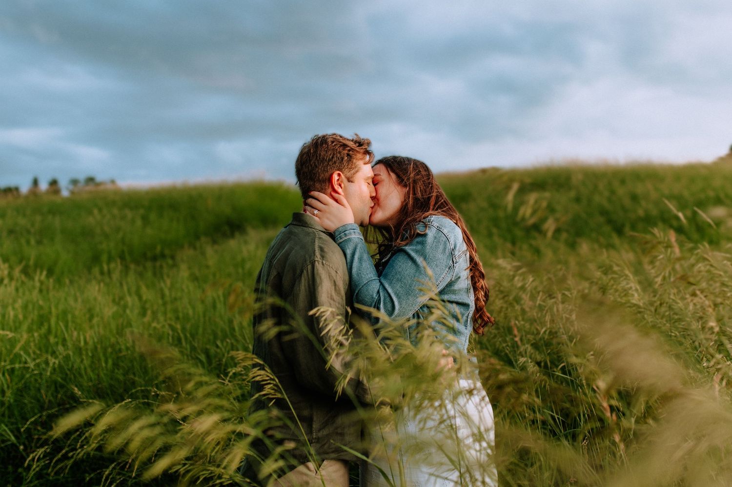 Couple sharing a romantic kiss in a lush green field with tall grass and dramatic cloudy sky backdrop.