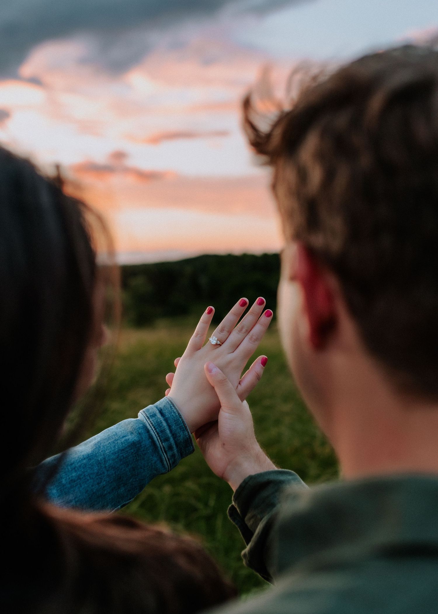 Couple holding hands at sunset, showing off an engagement ring with pink nails against a colorful sky.