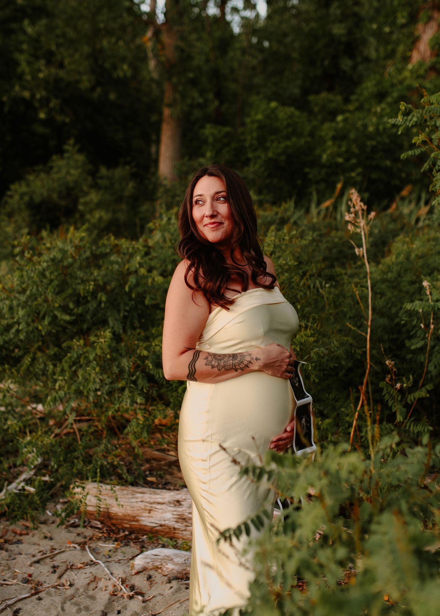Pregnant woman in yellow dress holding ultrasound photo outdoors among lush green trees during golden hour.