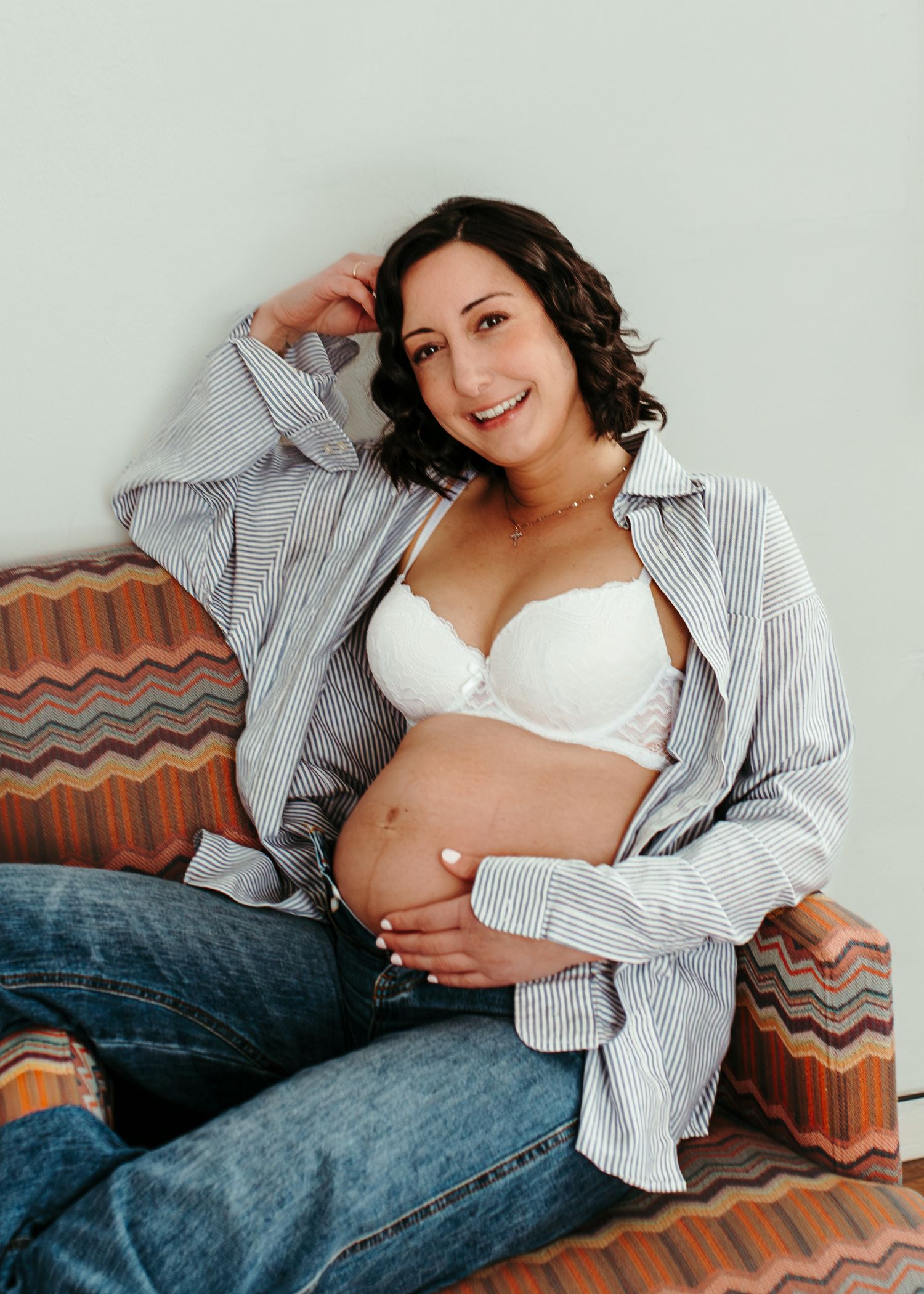 Smiling pregnant woman in white lace bra and open striped shirt sitting on colorful sofa cradling her baby bump.