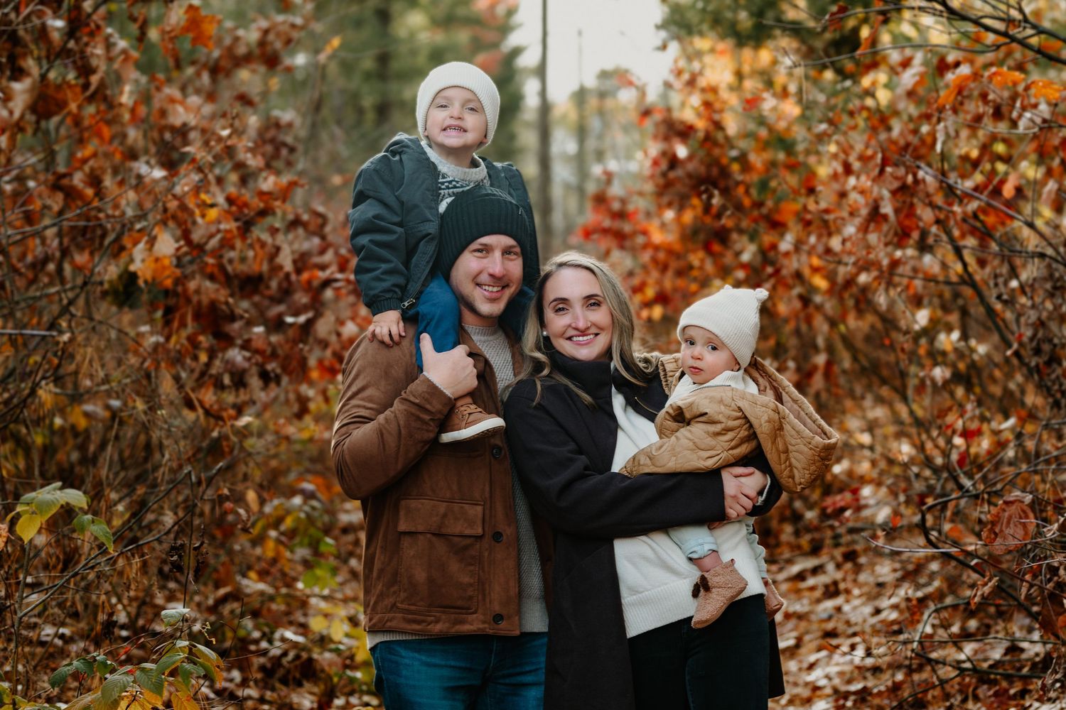 A couple and baby stand together among vibrant orange autumn foliage in a park setting.