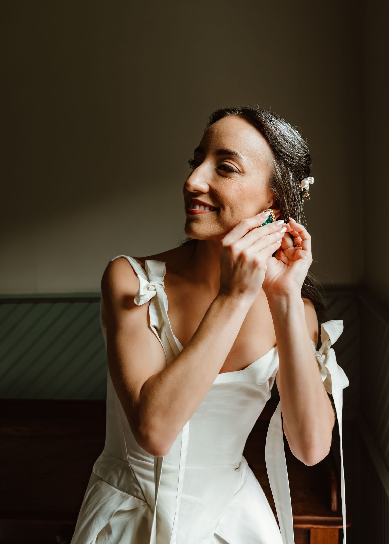 A joyful bride in a white dress puts on her earring while getting ready in a moody, dark room.