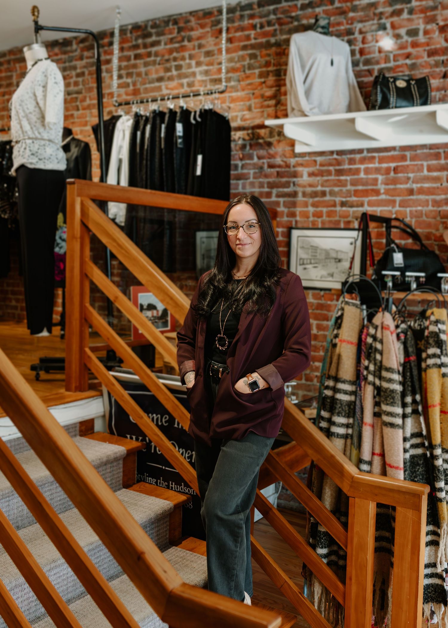 A person in a burgundy sweater and glasses sits on wooden stairs with exposed brick walls in a retail setting.