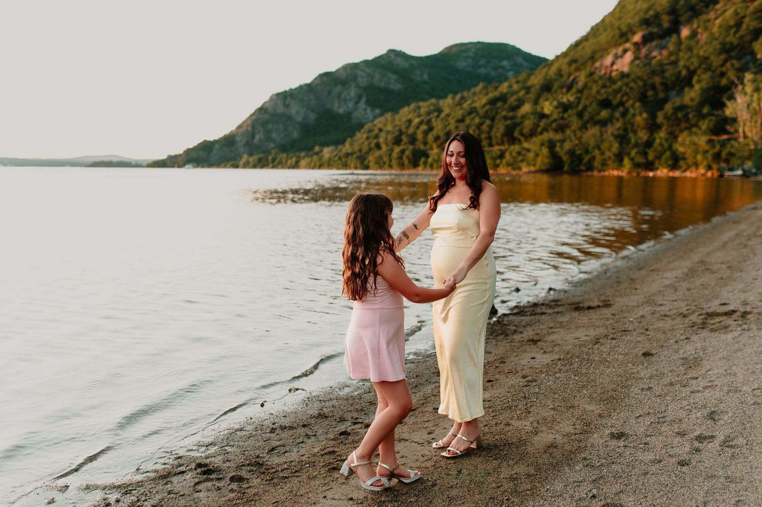 Pregnant woman in white dress holds hands with young girl on rocky beach with mountain lake backdrop.