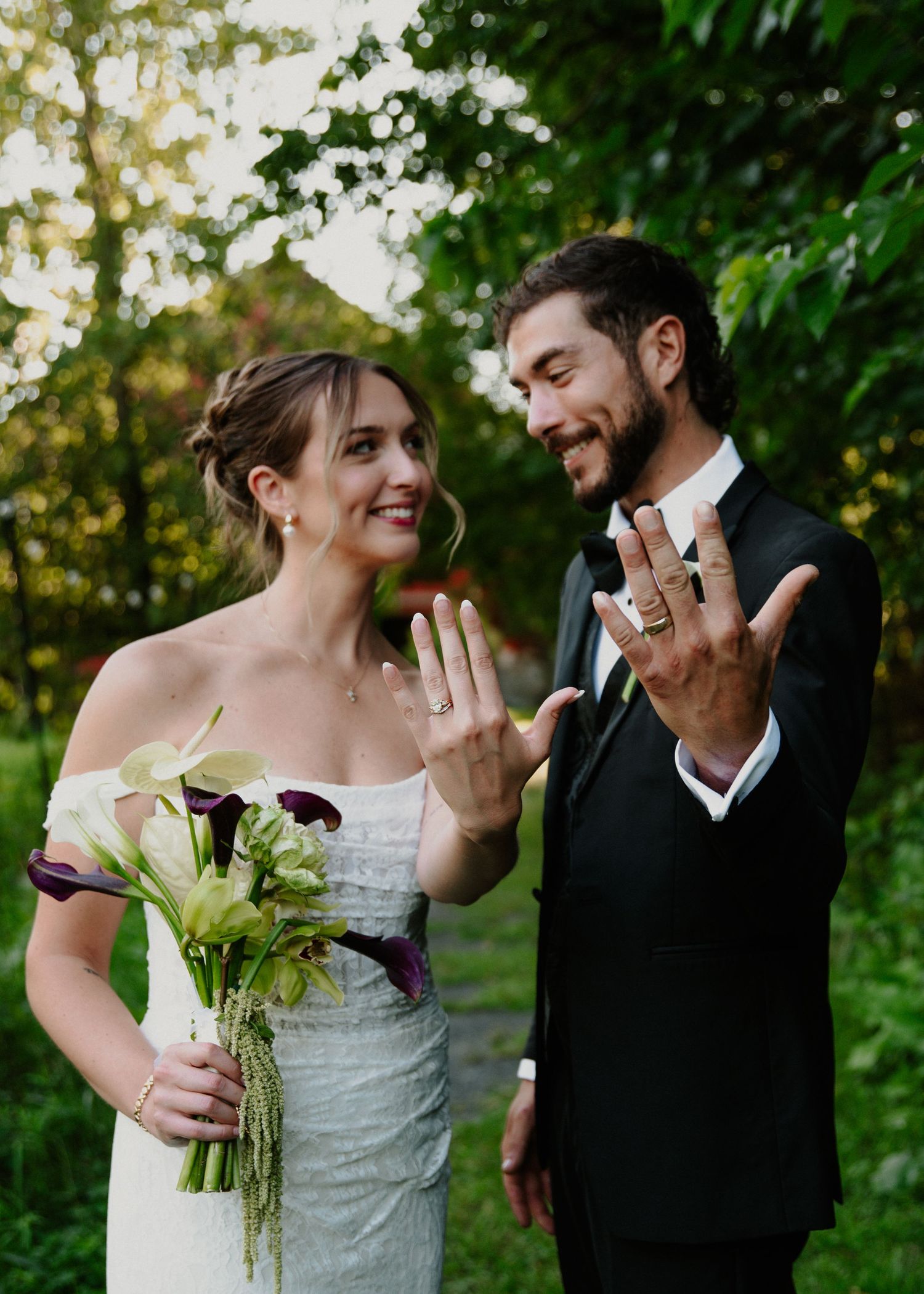 Newlyweds smiling and showing wedding rings, bride holding purple calla lily bouquet in white gown outdoors.