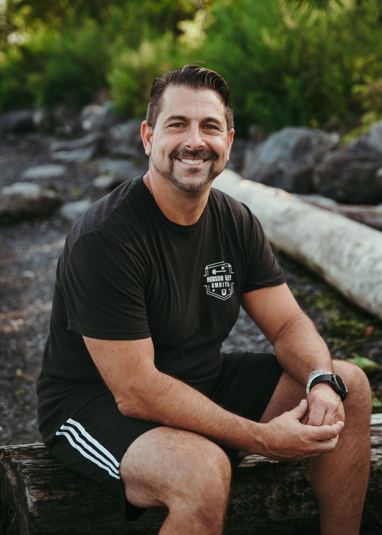 Person in black t-shirt with logo sits outdoors on rocks smiling at camera in natural setting.