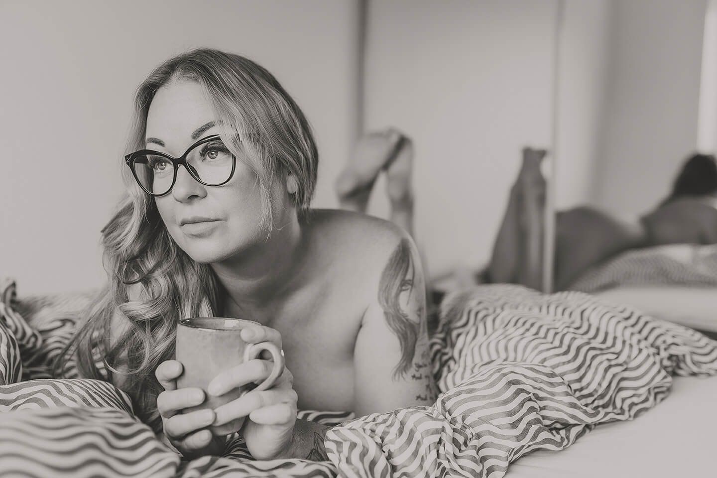 Boudoirfotografering i Aalborg – kvinde i sengen med briller og kaffekop, afslappet og hverdagsintimt udtryk.