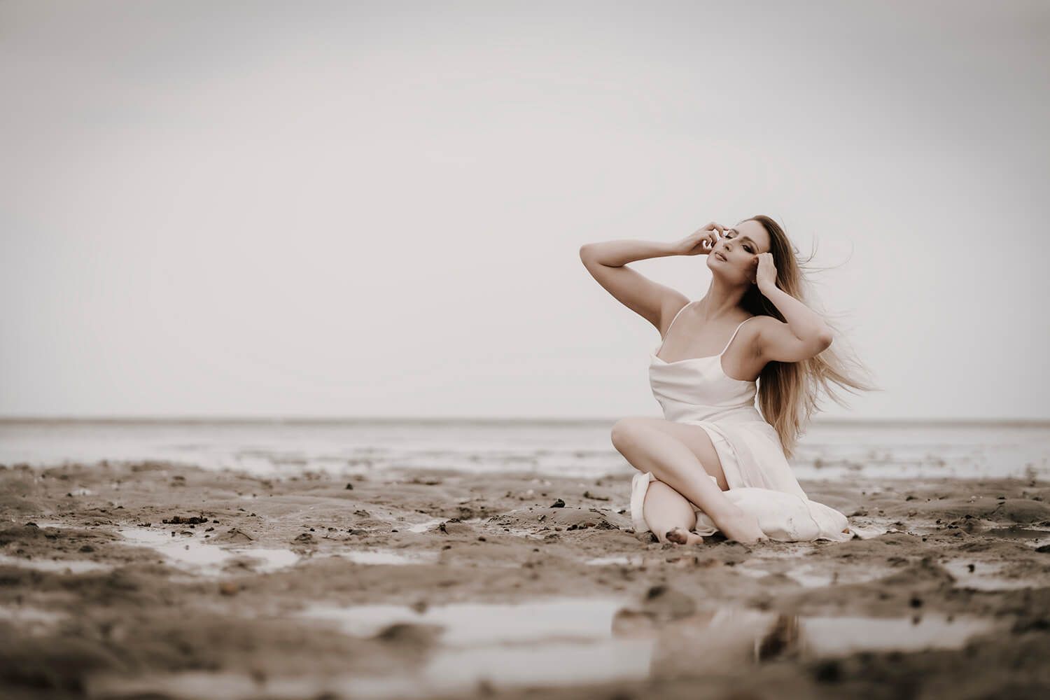 Modelfotografi på strand – kvinde i hvid kjole sidder med vind i håret i nordisk natur.
